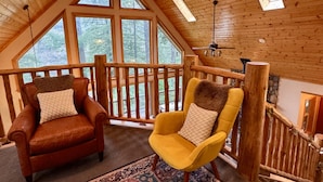 Ein gemütliches Schlafzimmer im Loftsstil mit schrägen Wänden, großen Fenstern und Blick ins Freie. Zwei Stühle mit Kissen und ein gemustertes Teppich sind auf dem Boden.