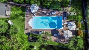 Une vue aérienne d’une piscine rectangulaire entourée d’une terrasse agrémentée de chaises longues et de parasols, le tout situé dans un paysage verdoyant luxuriant.