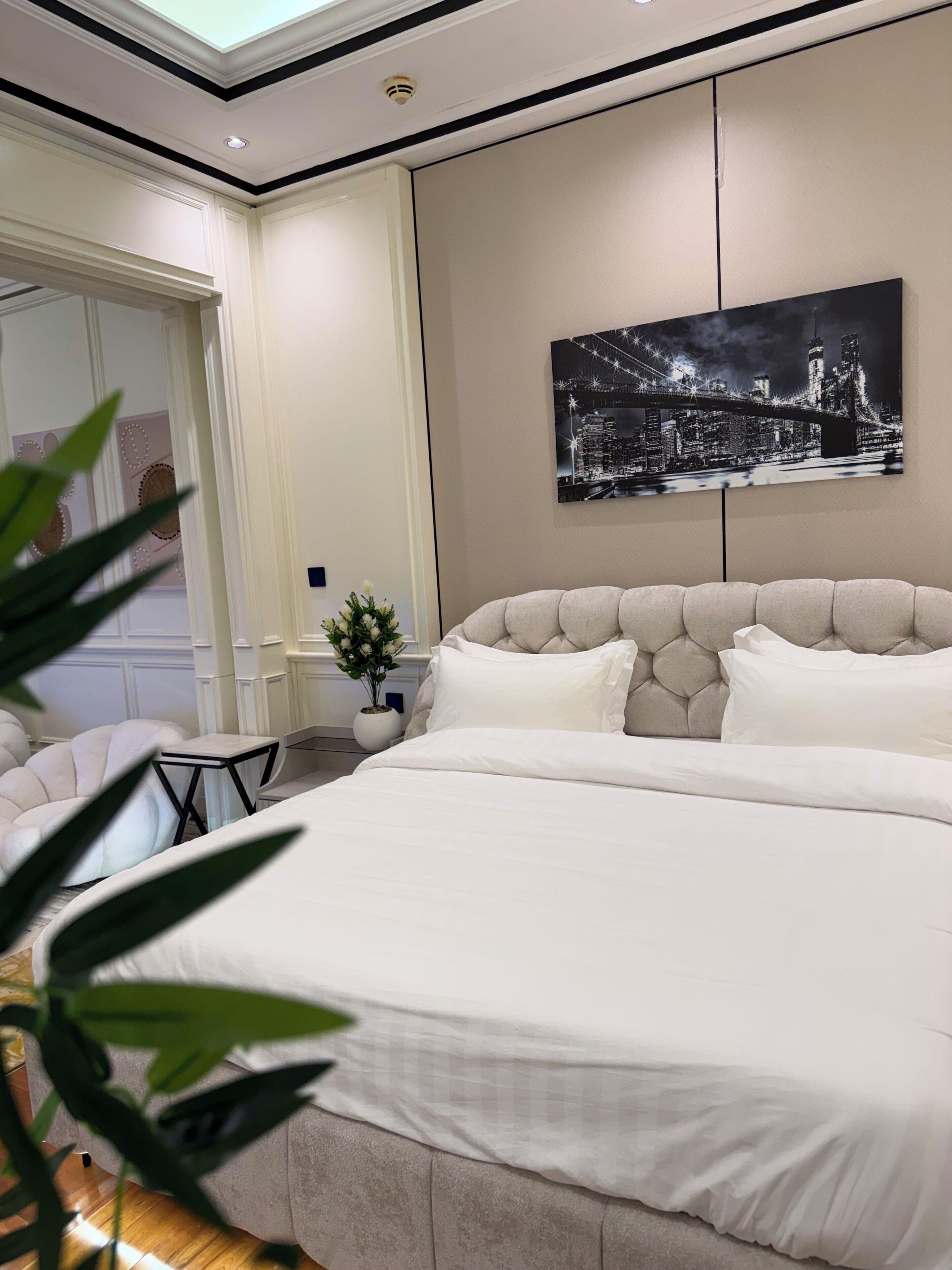 Ein Schlafzimmer mit einem großen Bett, einem Stadtbild an der Wand, einem kleinen Tisch mit einer Topfpflanze und einem Sessel.
