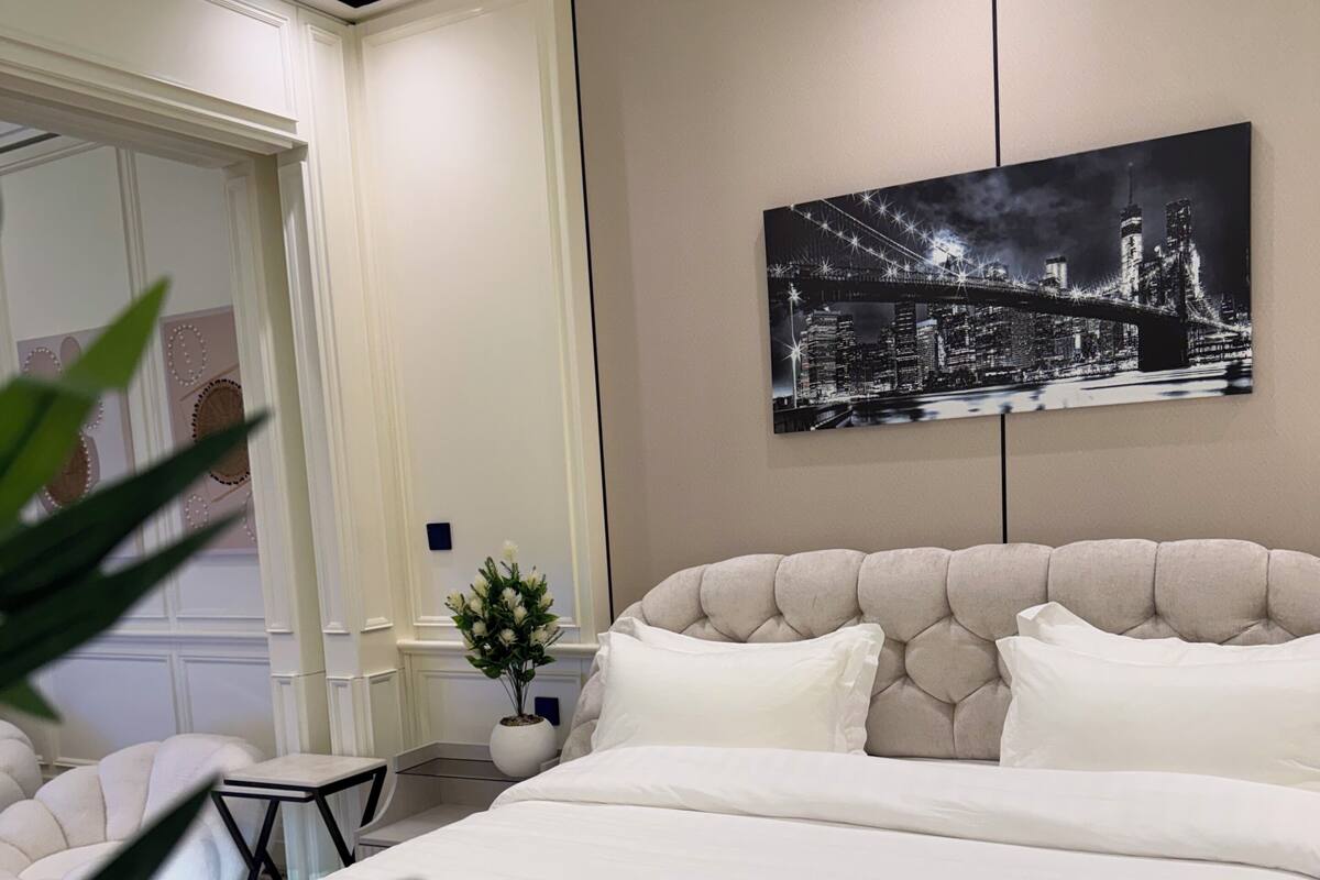 A bedroom with a large bed, a cityscape painting on the wall, a small table with a potted plant, and a chair.