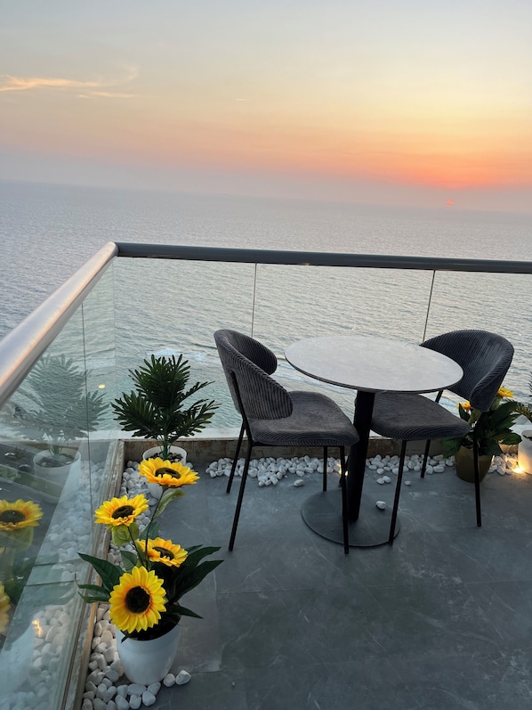 A balcony with a glass railing, a round table, two chairs, and potted sunflowers.