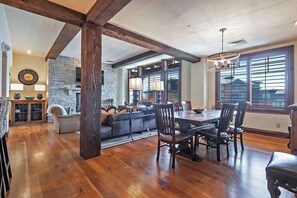 A spacious dining area with a large wooden table and chairs, a stone fireplace, and large windows with blinds.