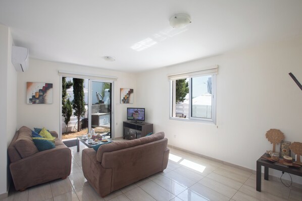 A living room with two sofas, a coffee table, and a television. There are windows with blinds, a ceiling light, and a small table with decorative items.