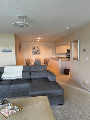 A living room with a gray sectional sofa, a coffee table, and a dining area with a table and chairs. There is a kitchen in the background with cabinets and appliances.