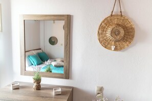 A bedroom with a wooden-framed mirror, a woven wall hanging, and a bedside table with a small plant and candle. - Emmanouela Apartment Matala (Matala)