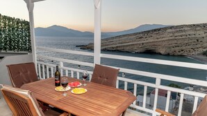 A wooden table with four chairs, a bottle of wine, and a glass of red wine, set on a balcony overlooking a coastal view with mountains in the background. - Emmanouela Apartment Matala (Matala)