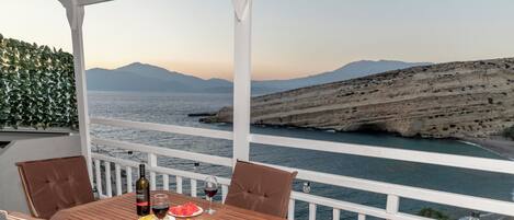 A wooden table with four chairs, a bottle of wine, and a glass of red wine, set on a balcony overlooking a coastal view with mountains in the background.