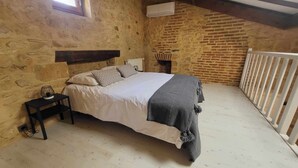 Et soveværelse med en stenvæg, trælofter, en seng med hvide sengetøj, et natbord med en lampe og et hvidt gelænder.