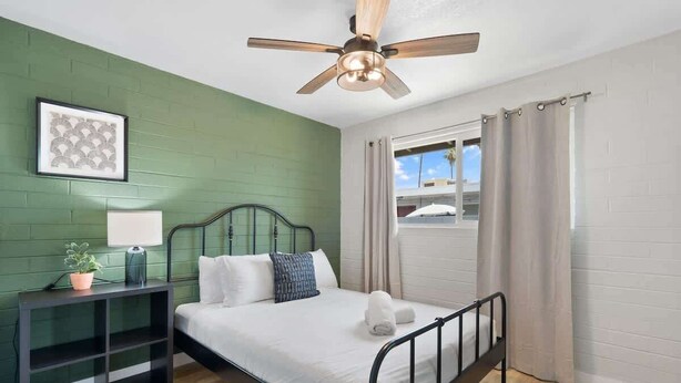 A room with a bed, a ceiling fan, a window with curtains, a bedside table with a lamp, and a framed picture on the wall.