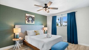 A hotel room with a bed, a ceiling fan, a window with curtains, and a small table with a lamp.