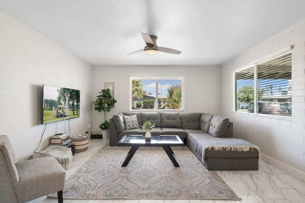 A living room with a sectional sofa, a glass coffee table, a ceiling fan, and a large window with blinds.