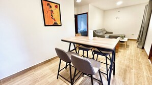 A modern living room with a wooden dining table, two bar stools, a grey sofa, and a small white side table.