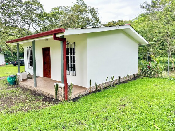 Una casa pequeña de un piso con fachada blanca y techo rojo. Cuenta con un porche con suelo de baldosas y un pequeño jardín con césped y plantas.