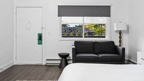 A modern hotel room with a bed, a dark gray sofa, a small side table, a lamp, and a window with a view of buildings and trees. - Peninsula One unit 18 Close to downtown and beach (Traverse City)