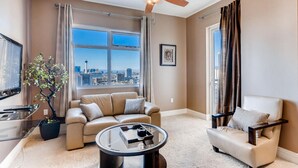 A modern living room with a sofa, armchair, and a coffee table. A ceiling fan is visible, and there is a large window with a view of a cityscape.