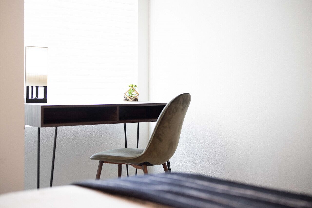 Ein Zimmer mit einem Bett, einem Schreibtisch und einem Stuhl. Es gibt ein Fenster mit Jalousien, eine kleine Pflanze auf dem Schreibtisch und eine Lampe auf dem Schreibtisch.