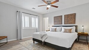 Une chambre à coucher comprenant un lit, un ventilateur de plafond, une fenêtre avec des rideaux et deux œuvres d’art murales.