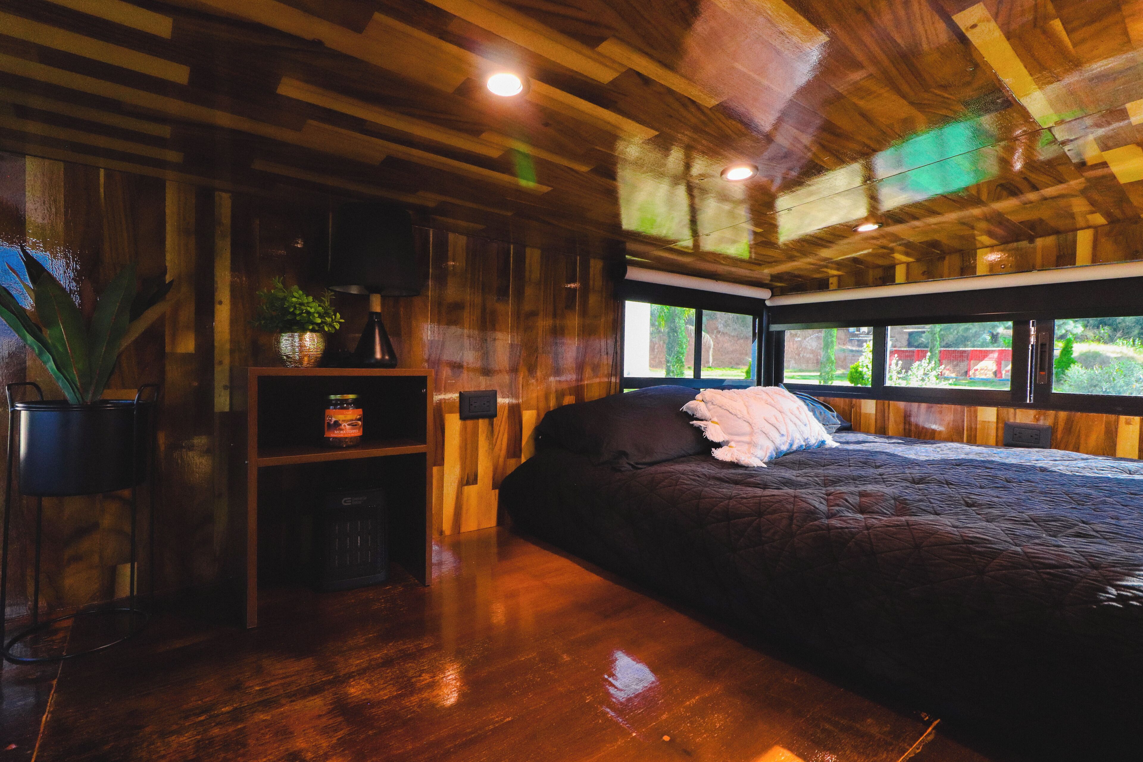 A bedroom with a wooden ceiling, a bed with a dark quilt, a small shelf with a plant and a jar, and a window with a view of greenery.