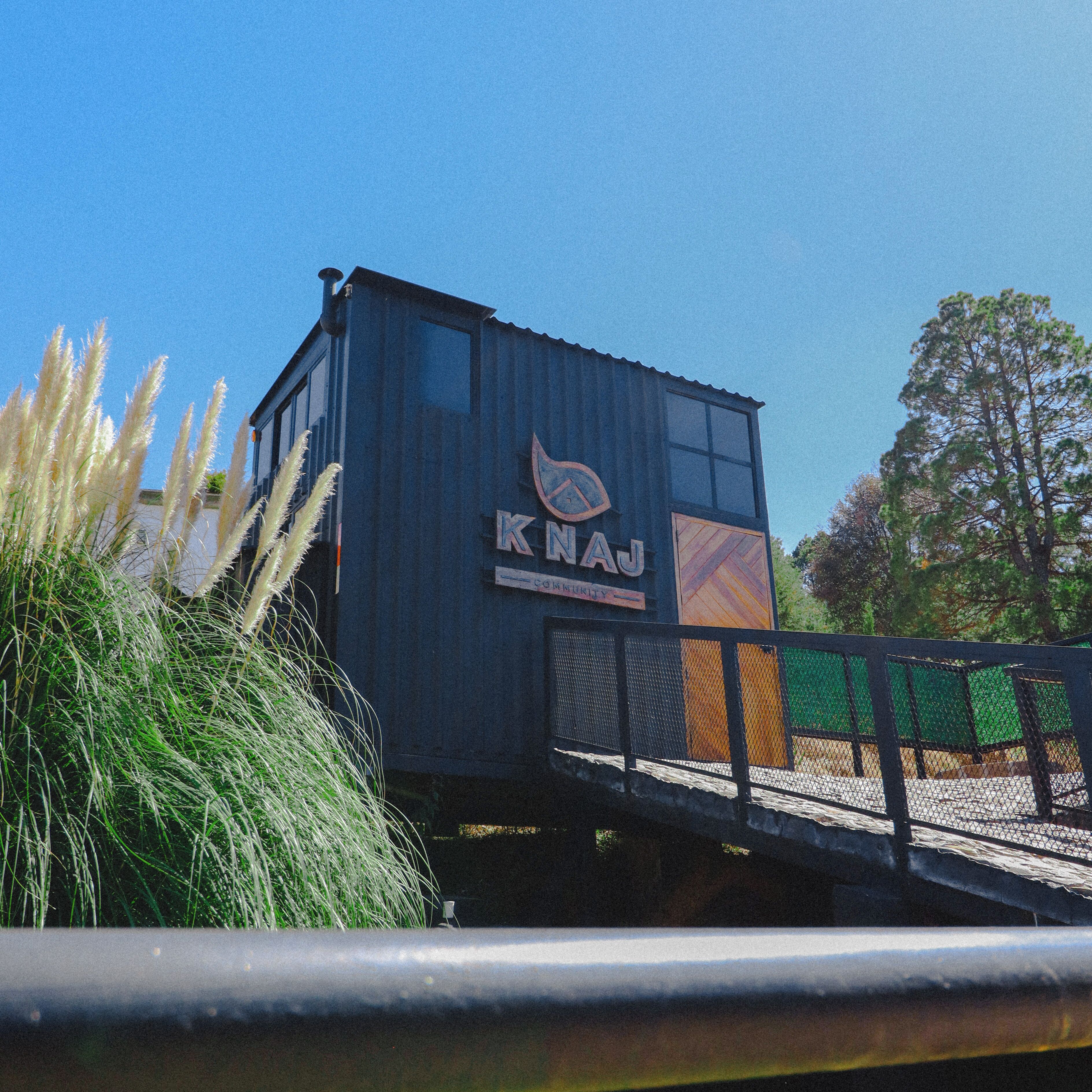 A small, dark-colored building with the sign "KNAJ" on its side, situated next to a green fence and surrounded by tall grass.
