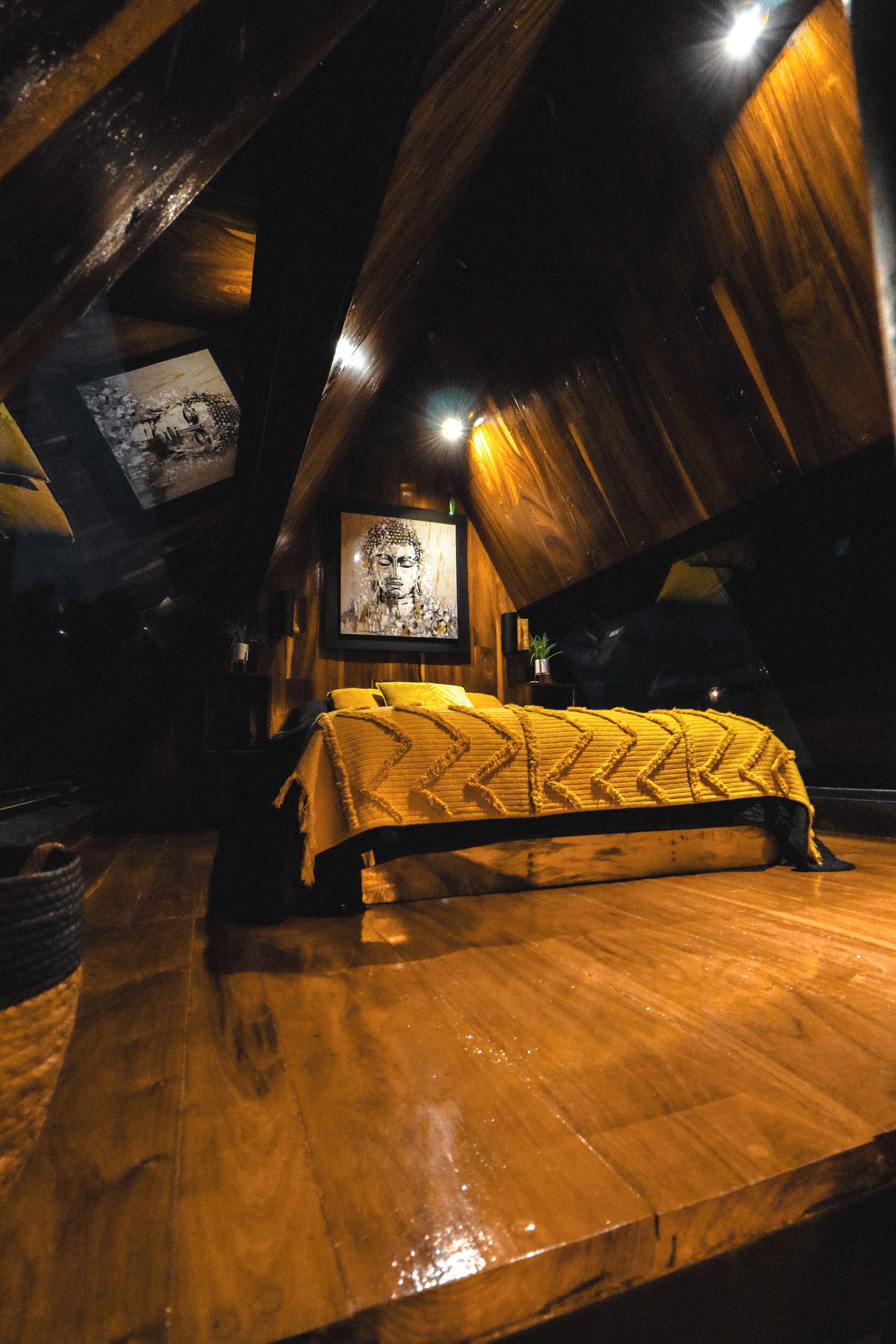 A cozy attic bedroom with a wooden floor, a bed with a patterned blanket, and a Buddha painting on the wall.