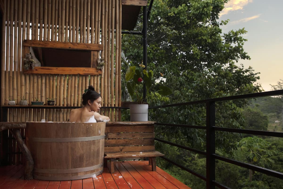 A wooden bathtub on a balcony with a bamboo wall and potted plants.