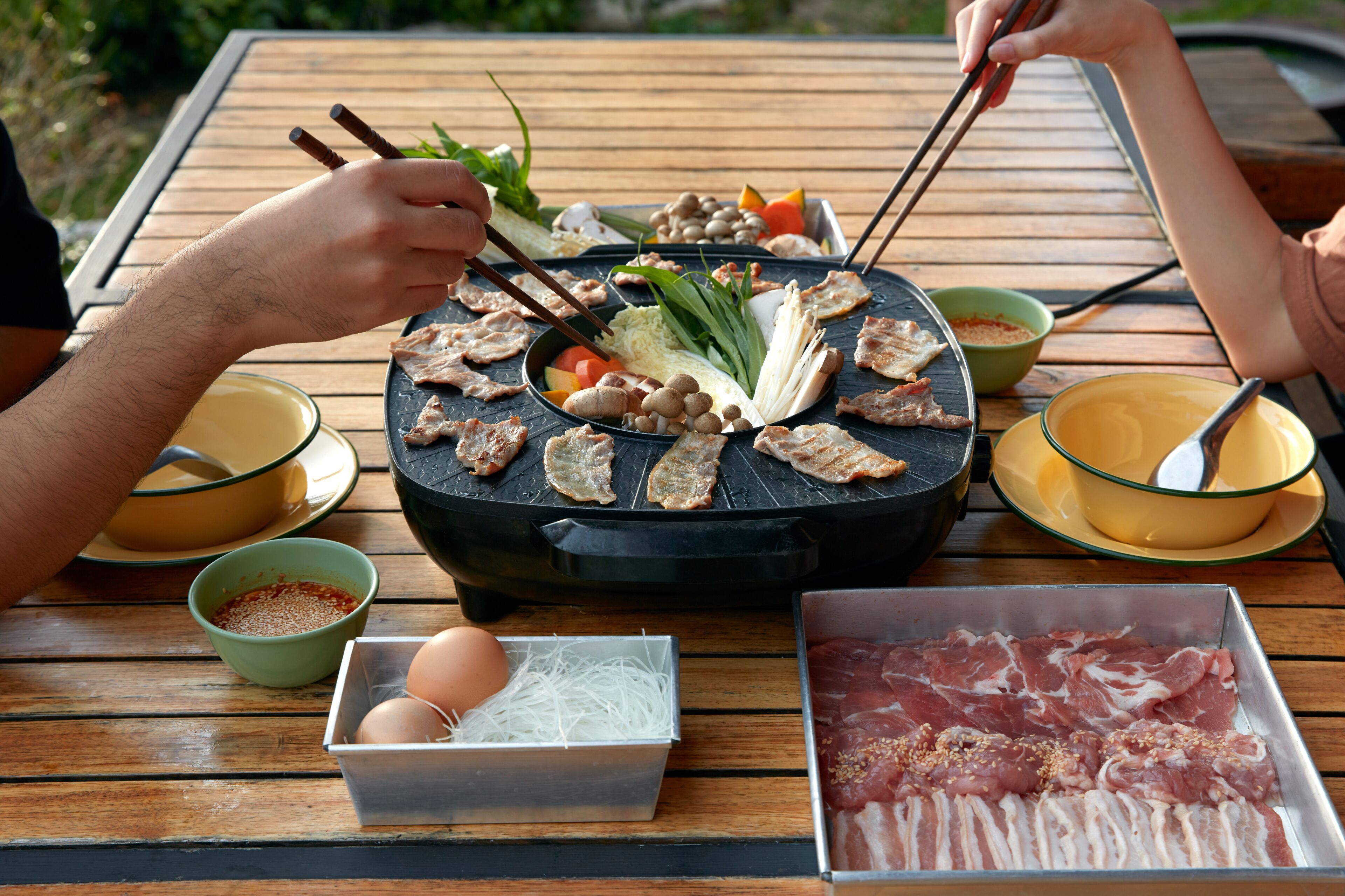 A wooden table with a black grill, various meats, vegetables, and condiments.