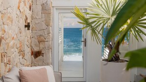 Habitació amb paret de pedra, porta blanca que dona a un balcó amb vista al mar, i una planta en test.