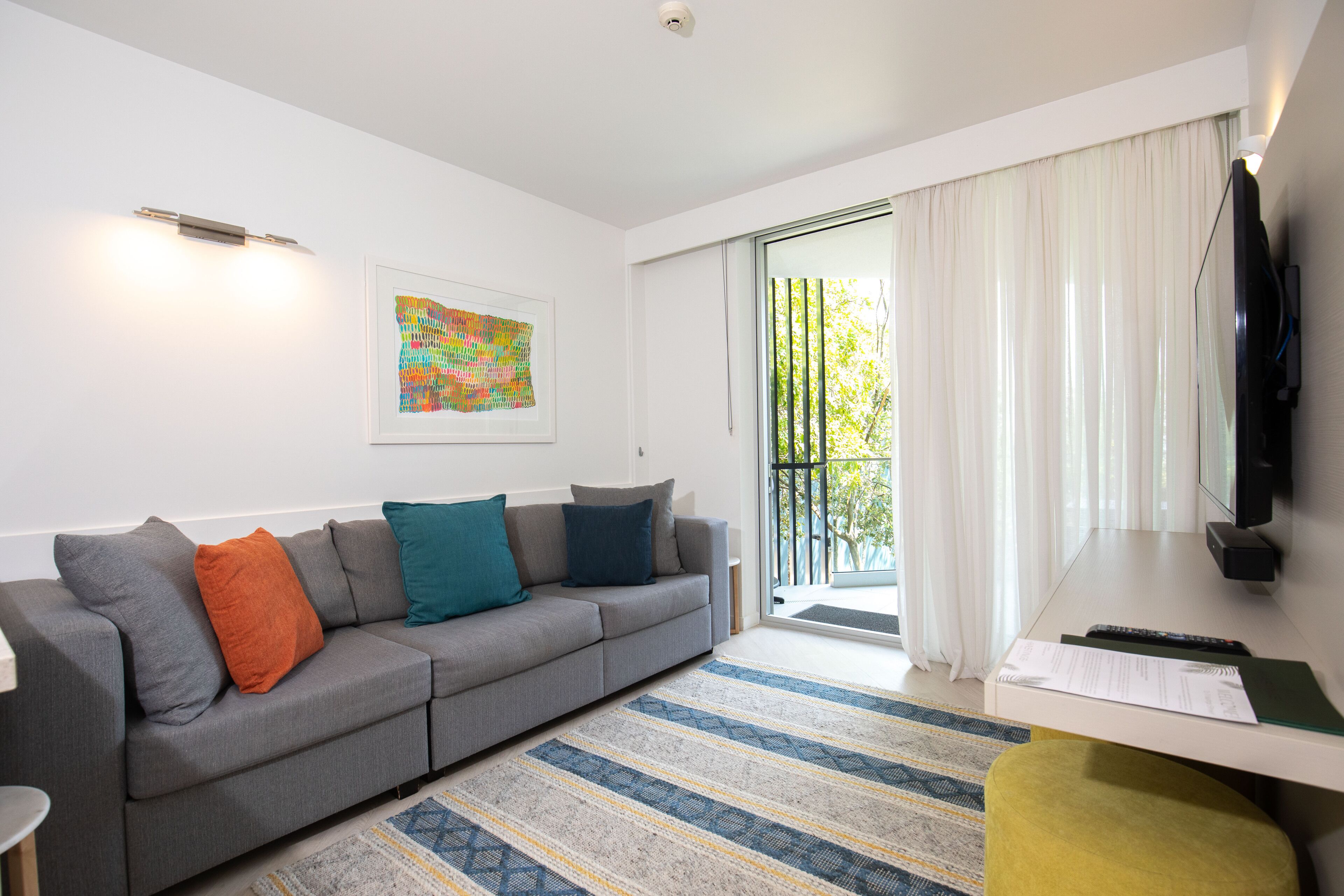 A modern living room with a grey sofa, colorful cushions, a striped rug, a flat-screen TV, and a sliding glass door leading to a balcony.