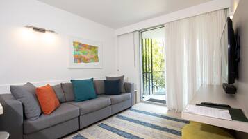 A modern living room with a grey sofa, colorful cushions, a striped rug, a flat-screen TV, and a sliding glass door leading to a balcony.