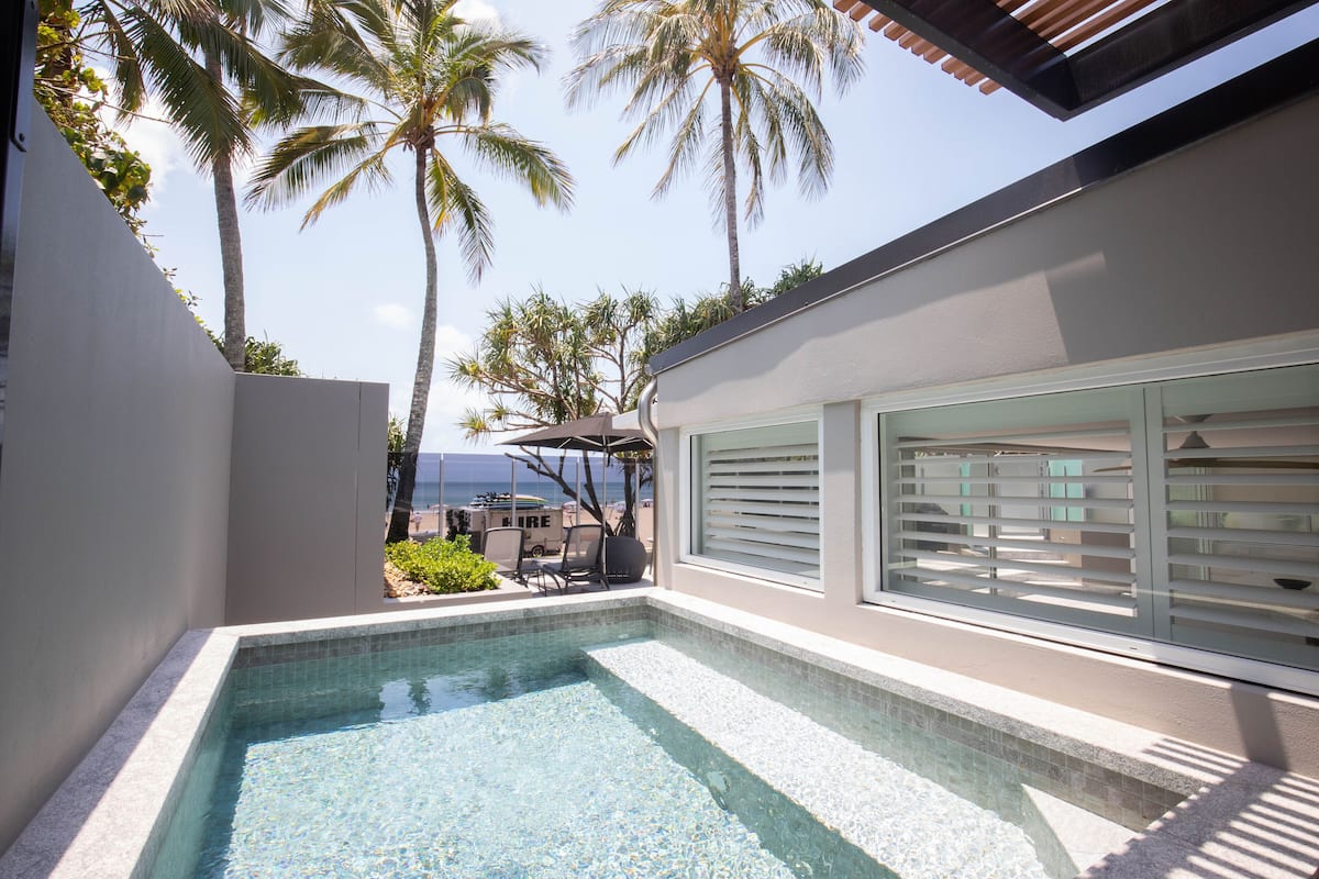 A modern outdoor swimming pool with clear blue water, surrounded by a white wall and palm trees.