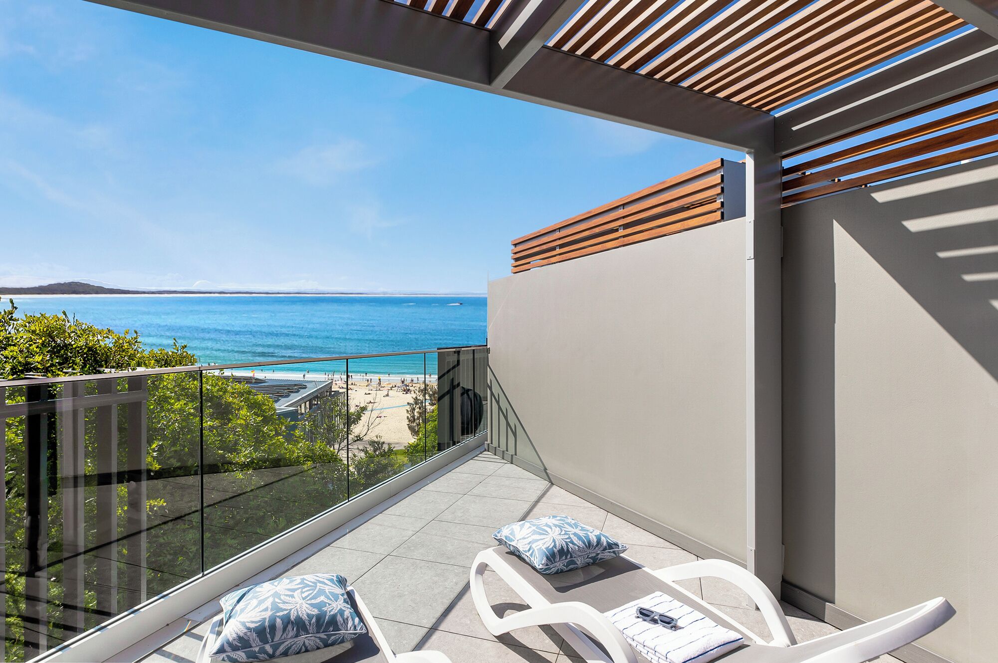 A balcony with a view of the sea, a glass railing, and two white lounge chairs with cushions.