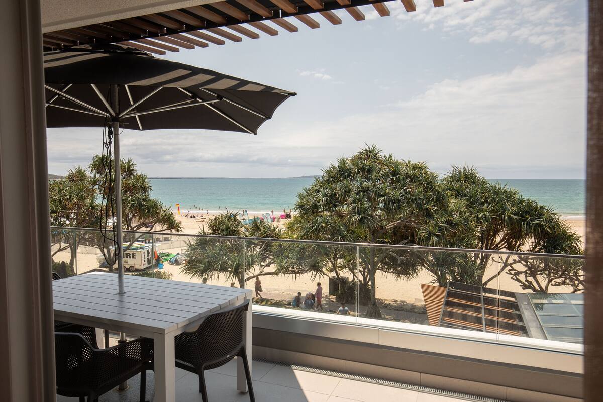 A balcony with a table and chairs, overlooking a beach with palm trees and the ocean.