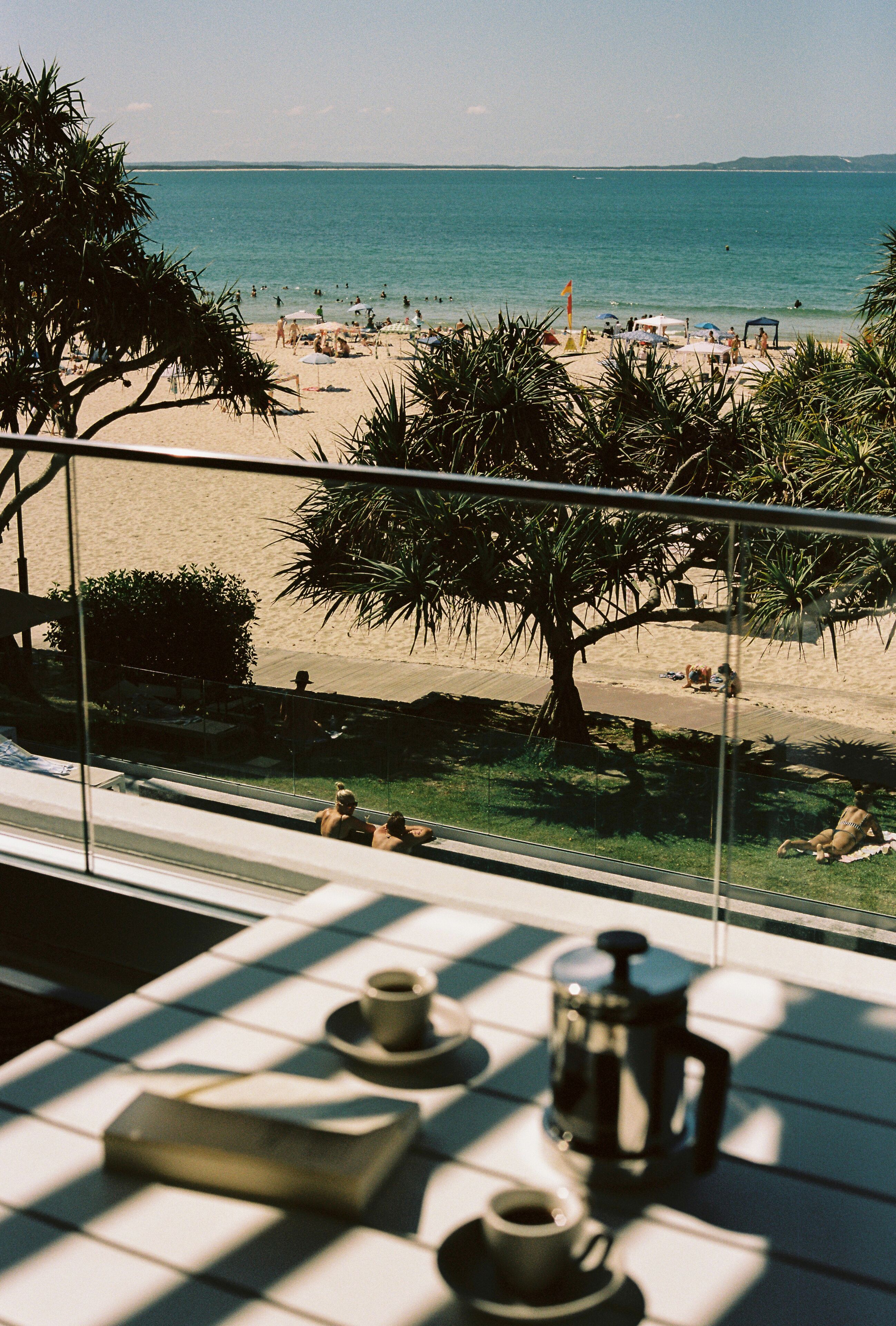 Un balcón con vistas a la playa, una cafetera y tazas sobre la mesa.