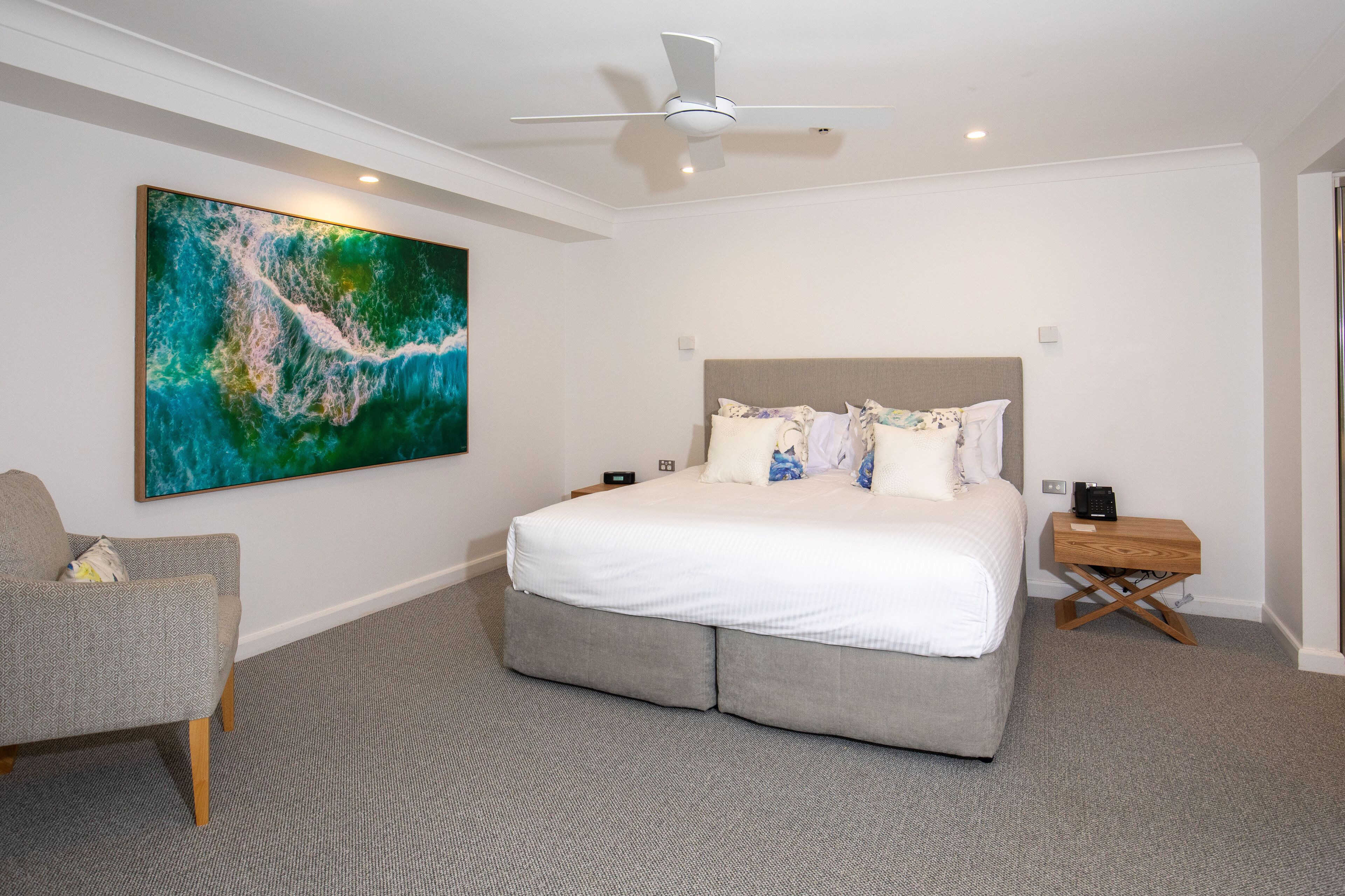 A bedroom with a large bed, a ceiling fan, a nightstand, a chair, and a large abstract painting on the wall.