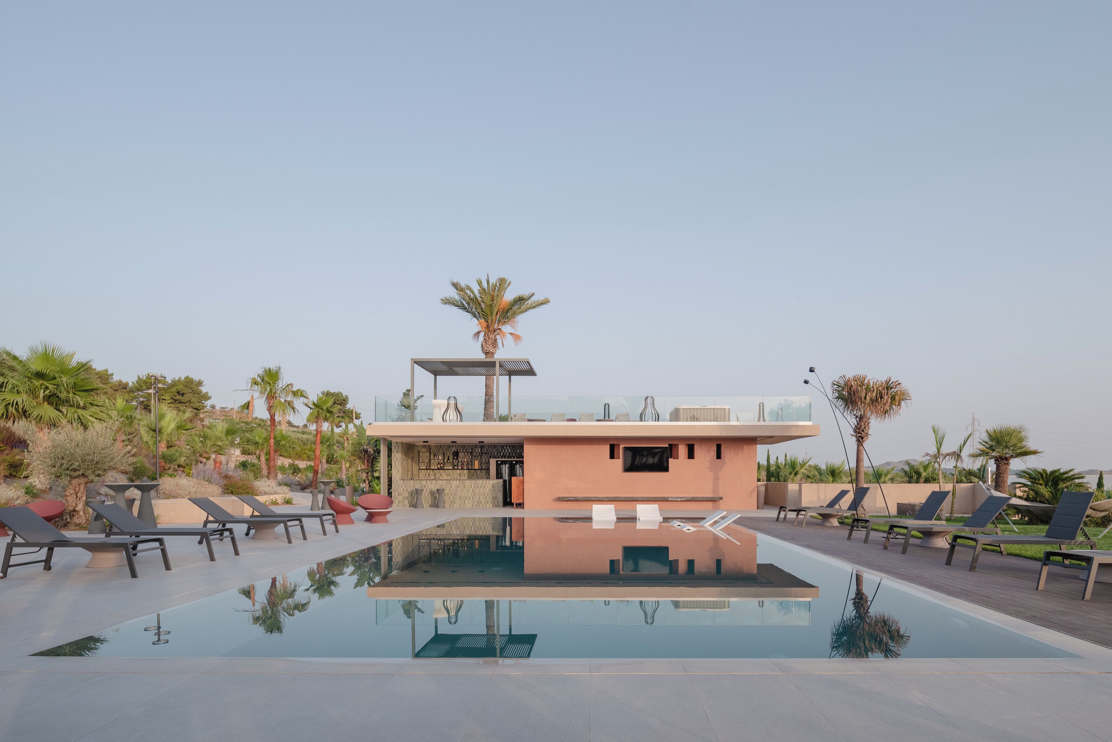 Un'area piscina moderna con poltrone prendisole, un piccolo edificio e palme.
