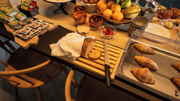 Un bufé de desayuno con pan, bollería, frutas y vasos de yogur.