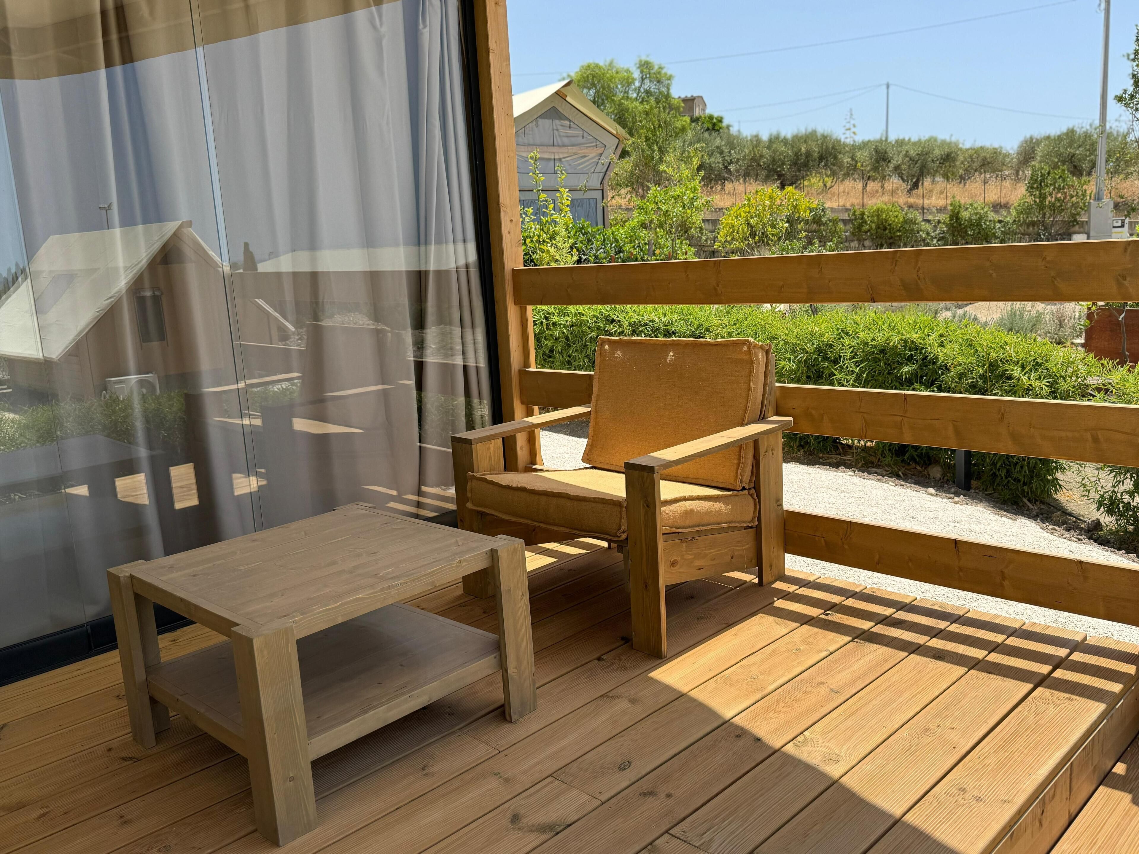A wooden deck with a chair and a small table, a wooden fence, and a view of greenery and buildings in the background.