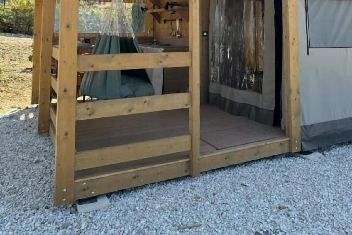 Eine Holzhütte mit geneigtem Dach, einer zur Einfahrt führenden Tür und einem kleinen, umzäunten Bereich mit einem hängenden Gegenstand.