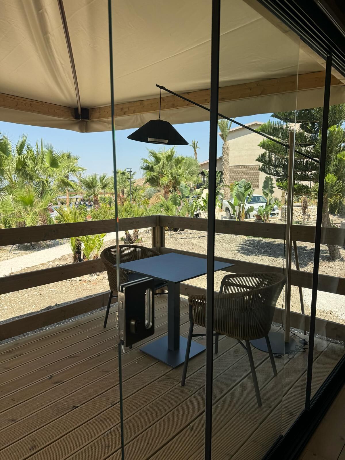 Une terrasse en bois avec une table et des chaises, une lampe suspendue, et une vue sur des palmiers et des immeubles.