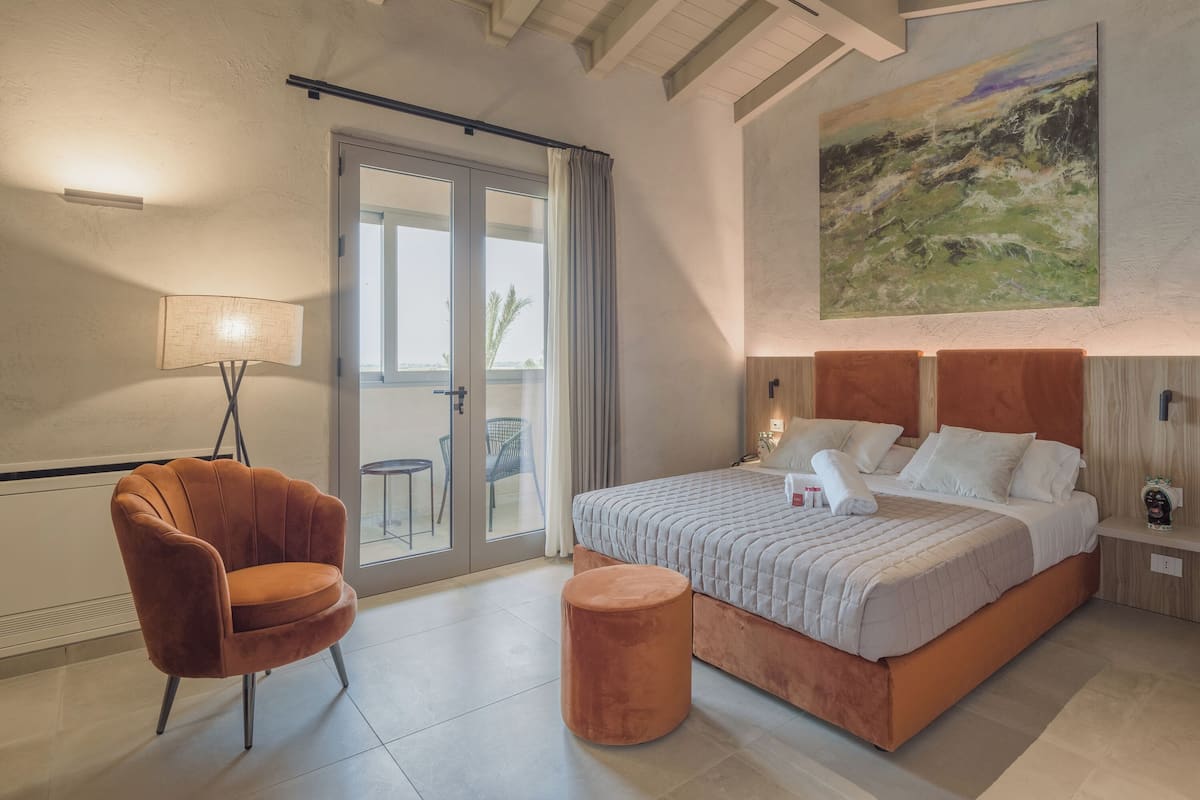 Ein Schlafzimmer mit Bett, einem Sessel, einem kleinen Hocker, einem Gemälde und einer Schiebetür zu einem Balkon.