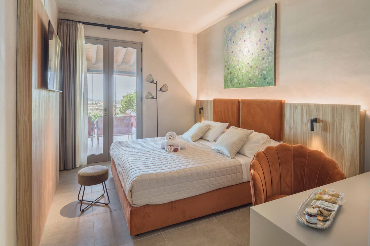 Ein Hotelzimmer mit Bett, einem kleinen Hocker, einem Tisch mit Snacks, einem Bild an der Wand und Blick durch die Tür.
