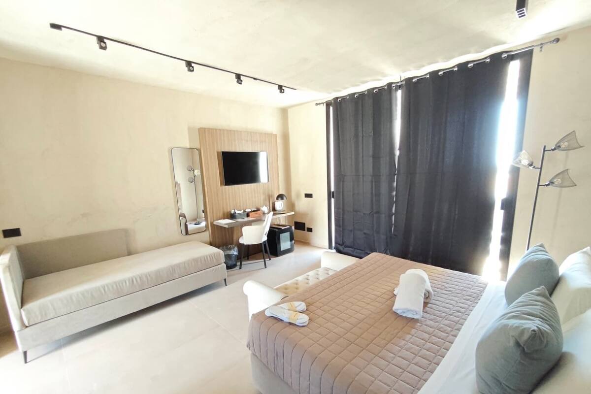 Ein Hotelzimmer mit Bett, einer Couch, einem Schreibtisch, einem Spiegel, einem Fernseher und einem großen Fenster mit Vorhängen.