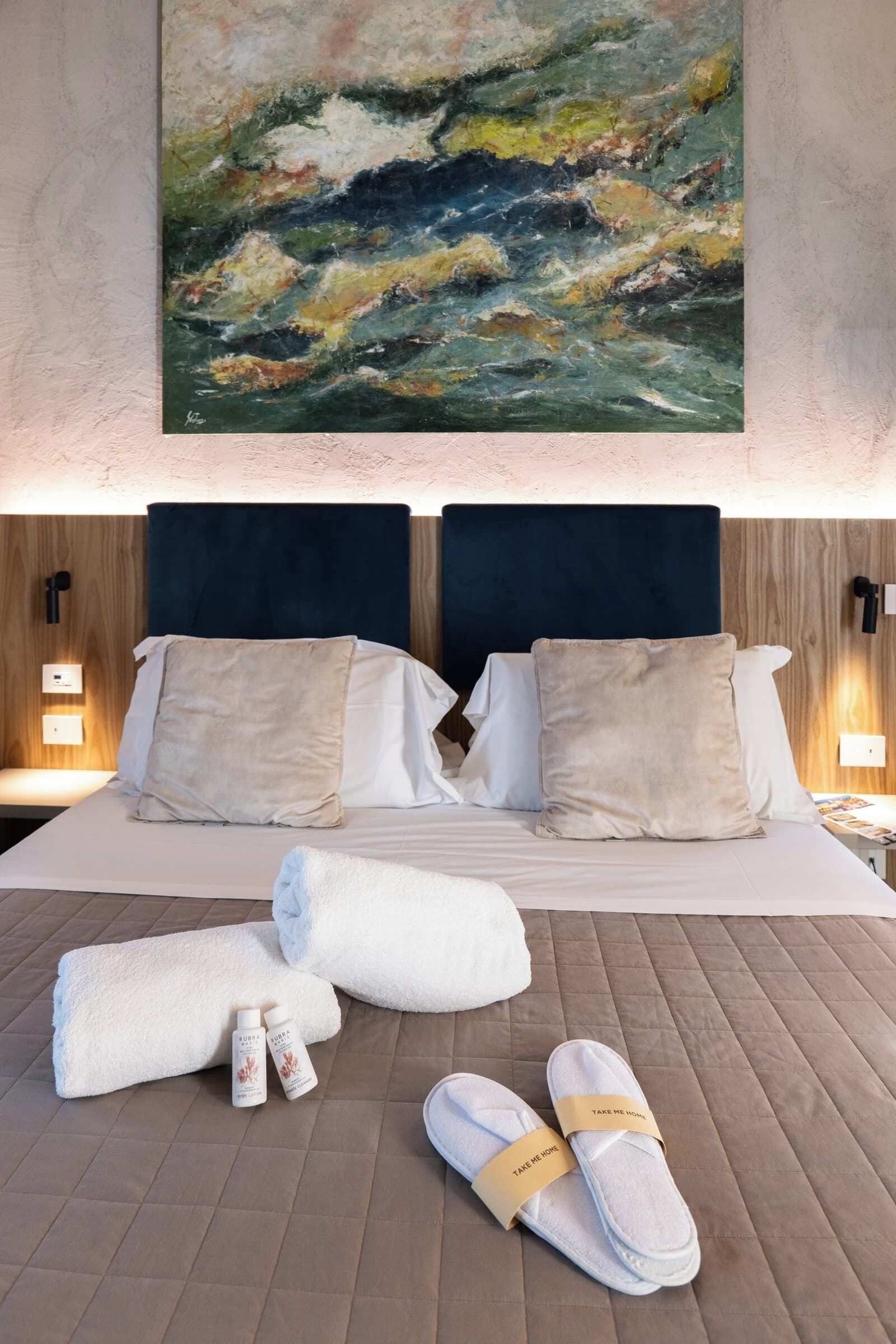A hotel room with a bed, pillows, towels, slippers, and a painting on the wall.