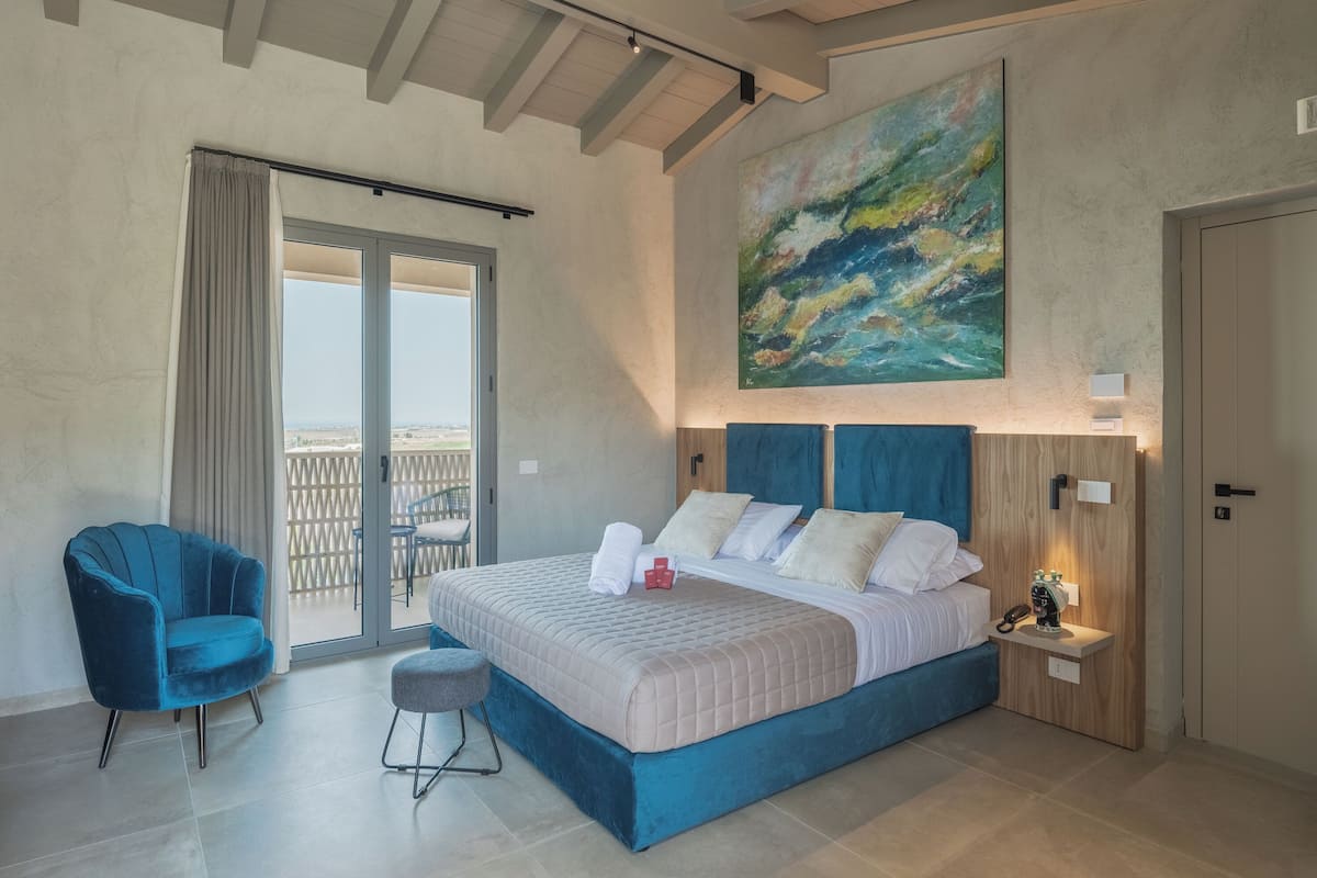 Ein Schlafzimmer mit einem großen Bett, einem blauen Sessel, einem kleinen Hocker, einem Nachttisch mit Telefon und einem Bild an der Wand.