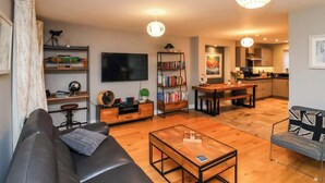 A modern living room with a sofa, coffee table, and a TV. There is a bookshelf, a dining area with a table and chairs, and a kitchen in the background. - The Beach Retreat (Filey)