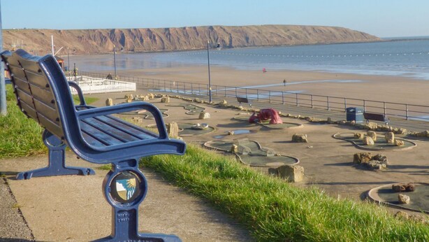 A bench with a circular emblem, a paved walkway, a rocky beach area, and a view of the sea and cliffs. - The Beach Retreat (Filey)