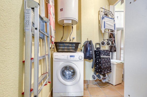 A compact laundry room with a front-loading washing machine, a stacked washer and dryer unit, a vacuum cleaner, and a small cart with a stroller.