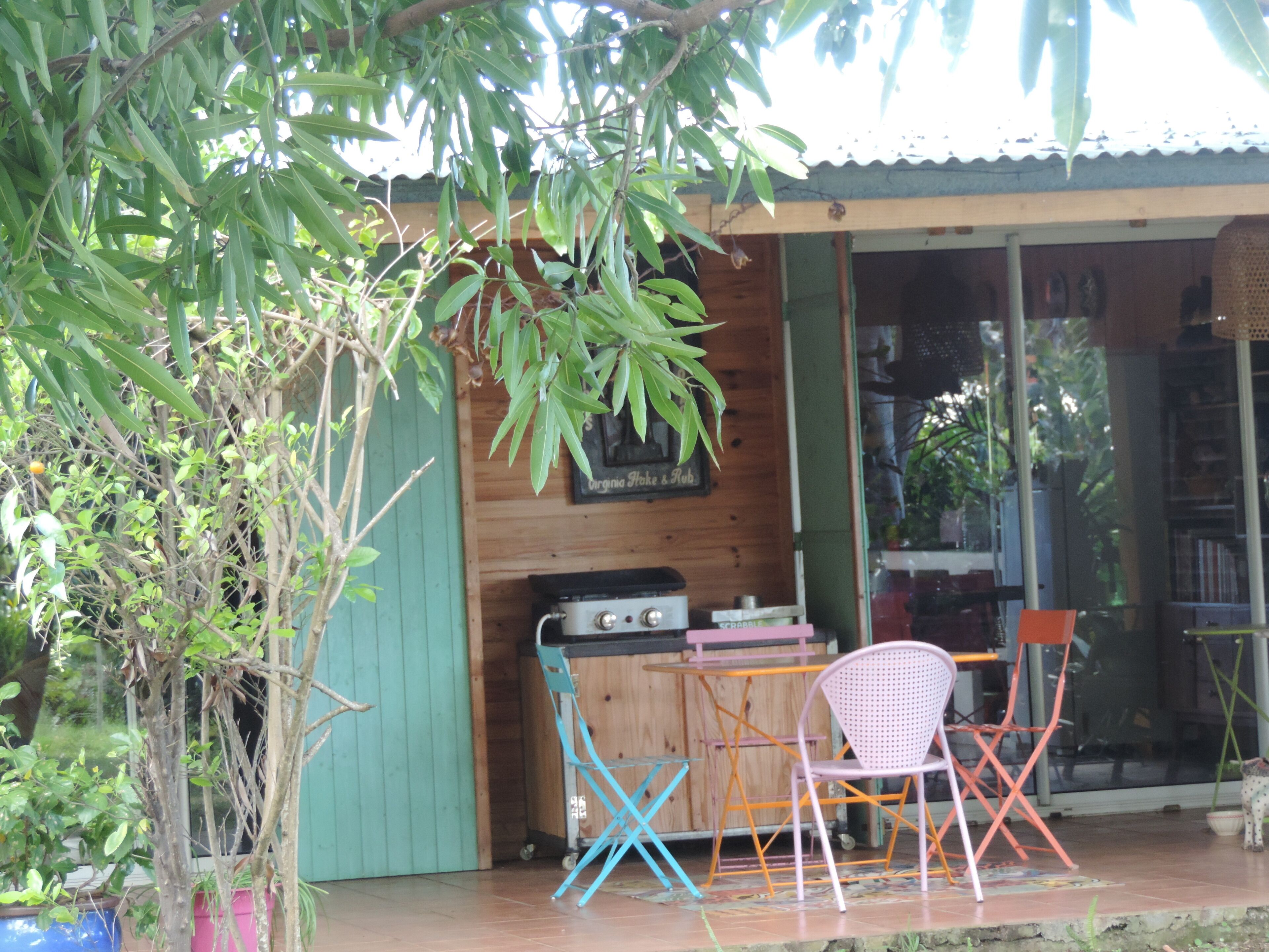 Une cabane en bois avec un petit espace extérieur aménagé pour s’asseoir, un barbecue et une fenêtre donnant sur la verdure.