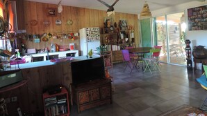 Amplia sala de estar con piso de madera, una mesa de comedor, sillas de colores, un área de cocina con refrigerador y un ventilador de techo.