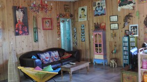 Una sala de estar acogedora con un muro de madera, un sofá negro con cojines de colores, un sillón amarillo, una mesa de centro de madera y varias decoraciones en la pared, incluyendo cuadros y una estantería.
